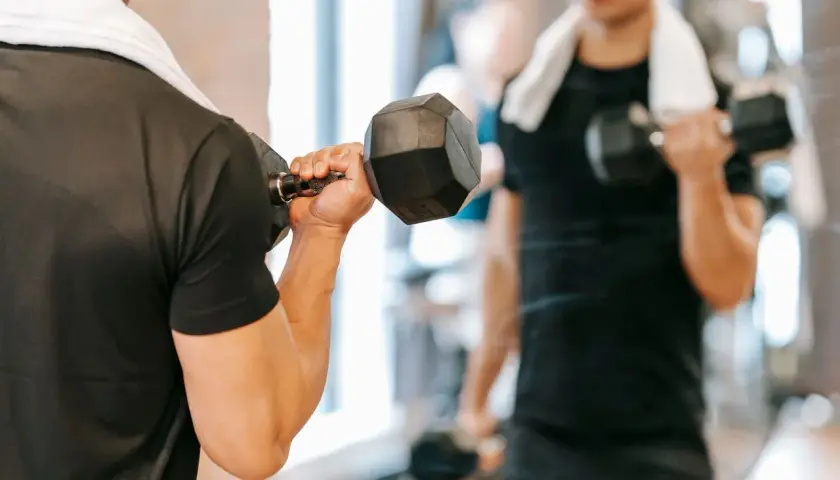 Mężczyzna ćwiczy biceps z hantlem, zaczynając dzień od treningu na czczo. W tle widać jego odbicie w lustrze.
