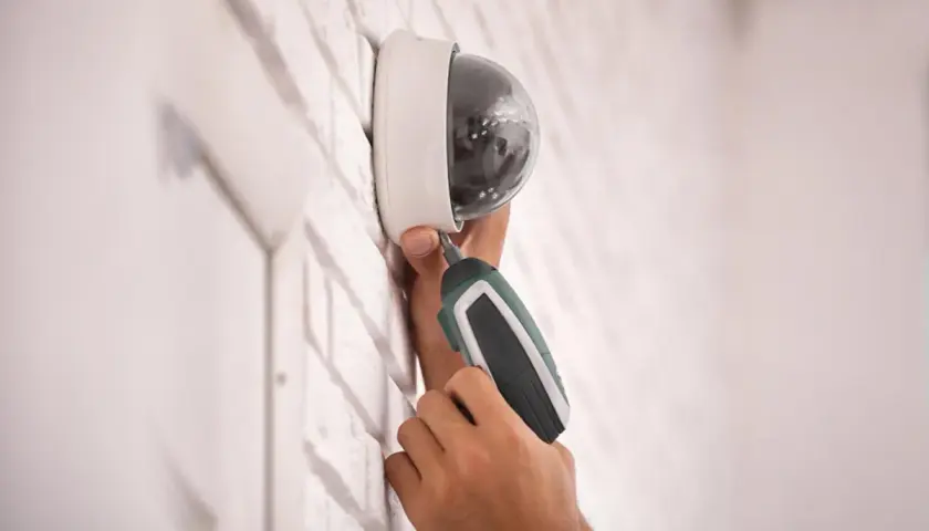 Installation d'un capteur infra rouge de surveillance, une main utilise un tournevis électrique pour fixer la caméra dôme sur un mur en briques blanches.