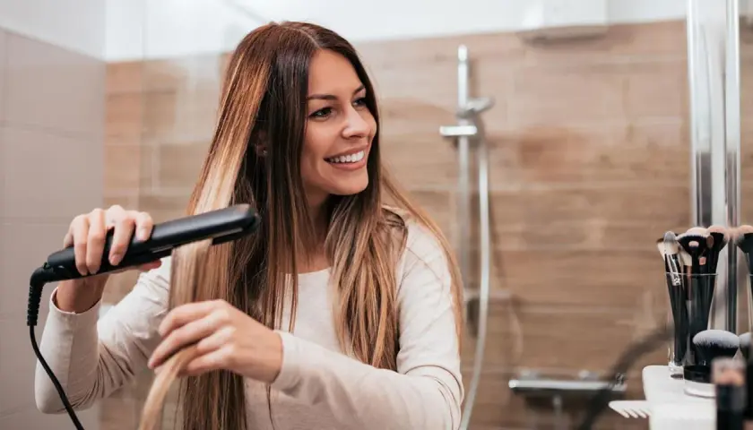 Mujer sonriendo mientras se alisa el pelo con una plancha, buscando el secreto de como alisar el pelo para siempre.