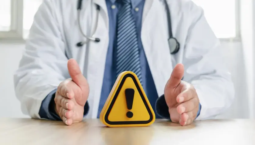 Medico con camice bianco e stetoscopio, mani aperte su un segnale di pericolo. Attenzione alle raccomandazioni ministeriali per la salute.