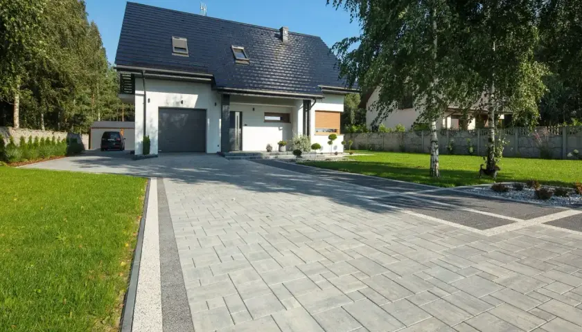 Nowoczesny podjazd przed domem z szarej kostki brukowej, otoczony zielonym trawnikiem i drzewami.