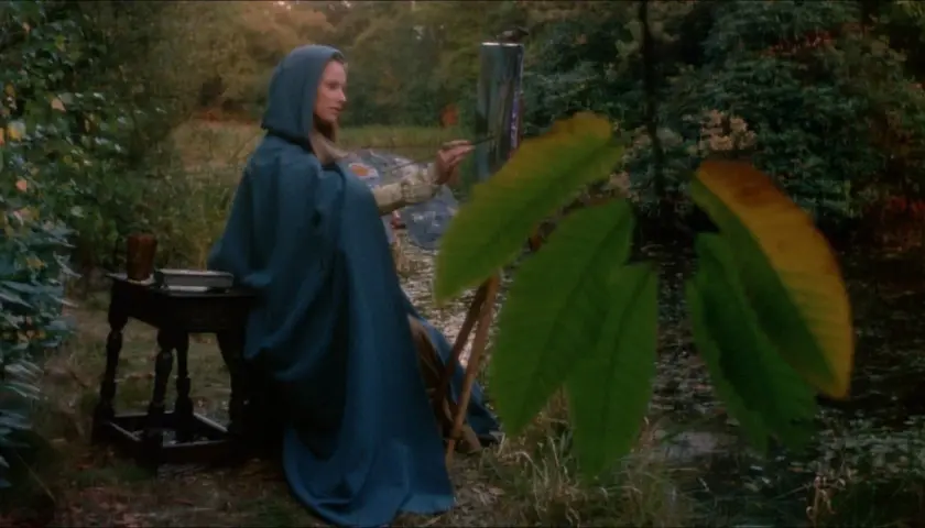 Essy Persson, klädd i en blå kappa, målar vid en sjö.