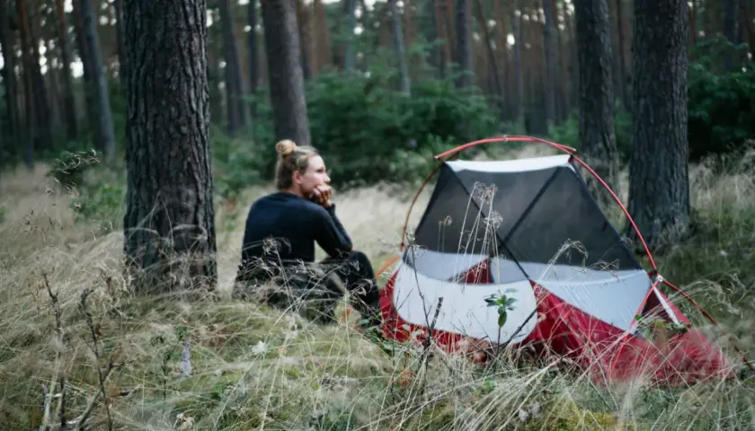 Ein Mann sitzt nachdenklich neben einem roten Zelt im Wald. Hier darf man im Wald zelten und die Natur genießen.