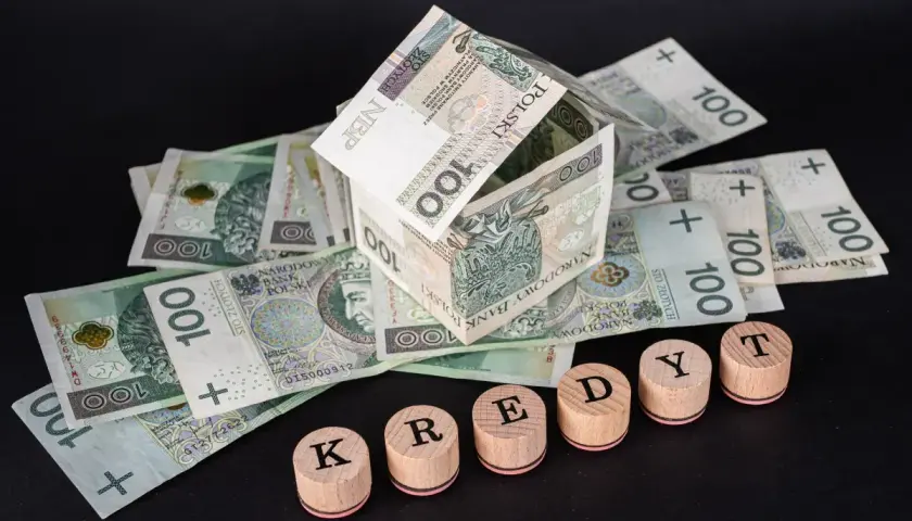 Dom z banknotów 100 zł, symbolizujący kredyt hipoteczny. Słowo "KREDYT" ułożone z drewnianych pieczątek.