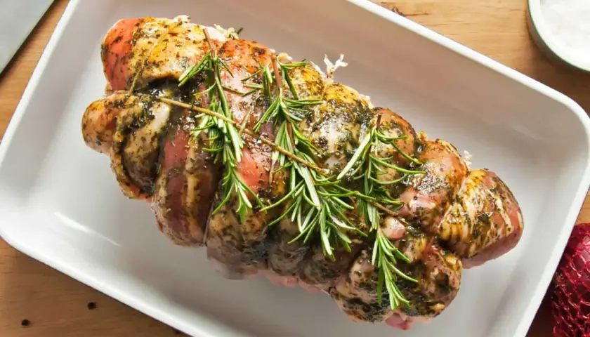 Un rôti de porc rosé, assaisonné et garni de romarin frais, prêt à être cuit dans un plat blanc.