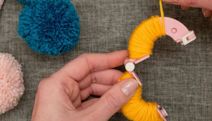 Hände wickeln gelbes Garn um eine Pompon-Schablone. Tolle Pompon-Ideen für dein nächstes Projekt!