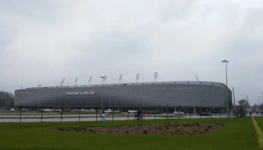 Nowoczesna Arena Lublin z napisem "ARENA LUBLIN" na fasadzie. Obok parking, a na pierwszym planie trawnik. Pojemność obiektu robi wrażenie.