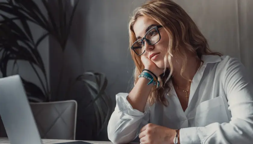 Kobieta w okularach opiera głowę na dłoni, patrząc zamyślona na laptopa.