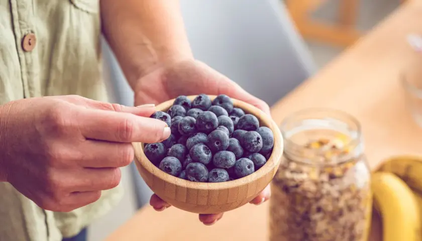 Hände halten eine Schale voller Blaubeeren, reich an Flavonoiden. Daneben stehen Müsli und Bananen – gesunde Flavonoide Lebensmittel für den Start in den Tag.