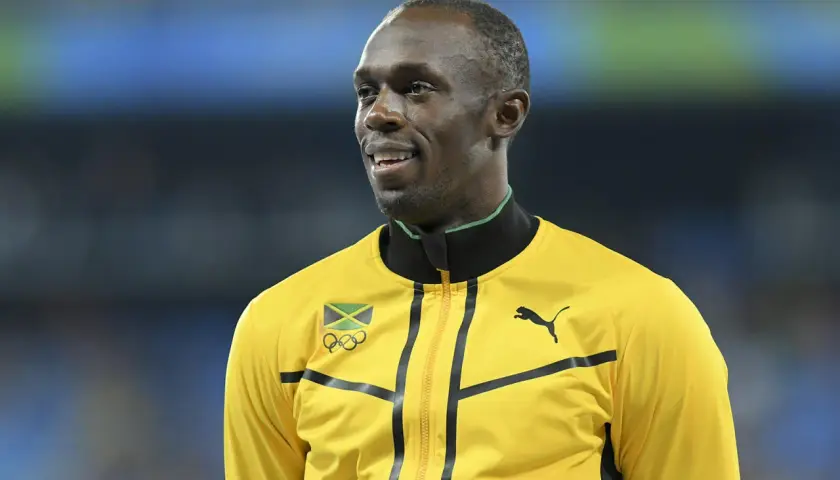 Usain Bolt w żółtej kurtce z flagą Jamajki i logo olimpijskim. Zastanawiasz się, jak szybko biega Usain Bolt?