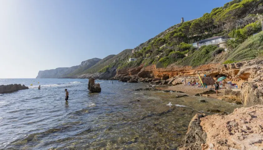 Gente disfruta del sol y el mar en una cala rocosa. Se ven casas y una torre en la **punta negra** de la colina verde.