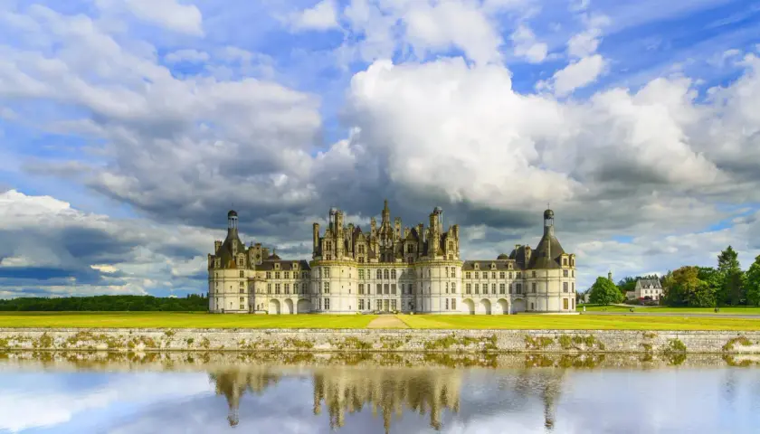 Zamek Chambord nad Loarą odbijający się w wodzie, pod burzliwym, błękitnym niebem.