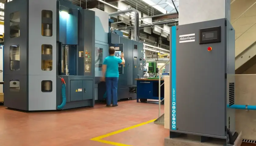 En una fábrica, un operario interactúa con una máquina industrial. A la derecha, un compresor Atlas Copco, que puede ser con aceite o sin aceite, espera su turno.