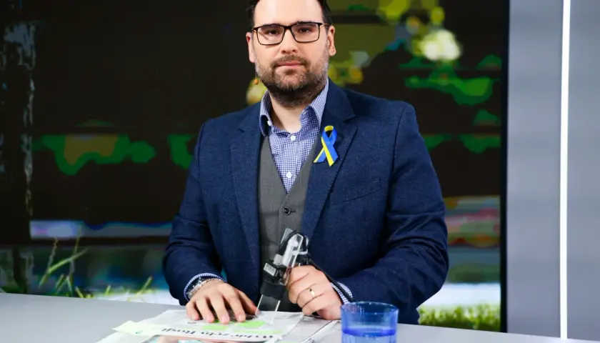 Michał Szajder, wiek nieznany, w garniturze z przypinką w barwach Ukrainy, siedzi przy stole z gazetami i szklanką wody.