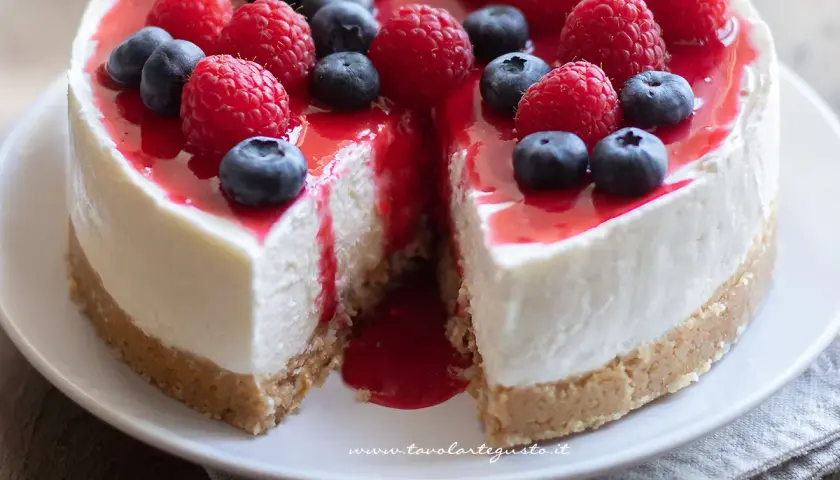 Cheesecake alla frutta con lamponi e mirtilli freschi, ricoperta da una glassa rossa lucida. Una fetta è stata tolta, rivelando la morbida crema.