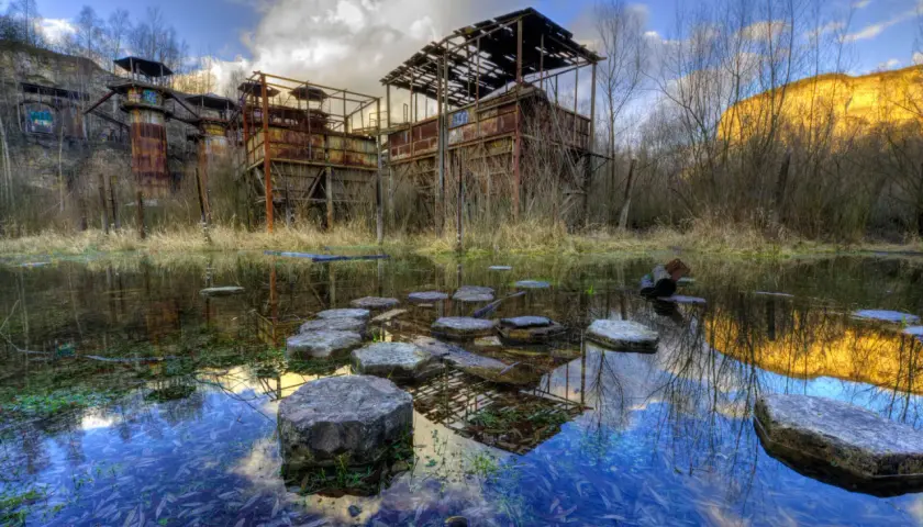 Zrujnowana fabryka, przypominająca opuszczoną "Fabbrica di Schindler", zardzewiałe konstrukcje i stawy odbijające niebo.