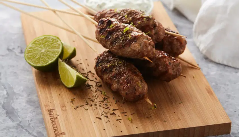 Brochettes d'agneau grillées, garnies d'herbes et de zeste de citron vert, servies avec des quartiers de citron vert et une sauce blanche.