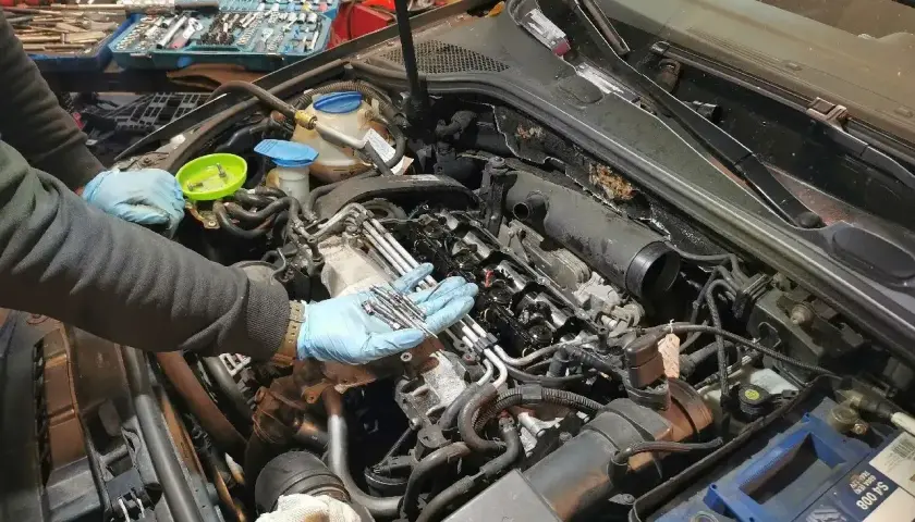 Mechanik w niebieskich rękawiczkach trzyma świece żarowe do wymiany. W tle skrzynka z narzędziami i silnik samochodu.