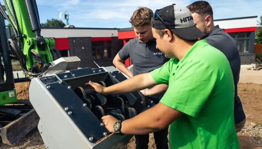 Trzech mężczyzn ogląda różne rodzaje łyżek do koparek, jeden z nich wskazuje na mechanizm.