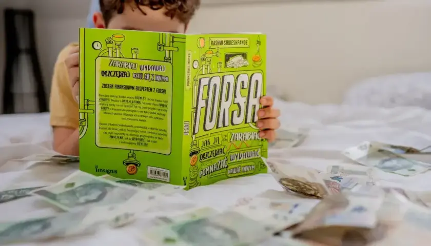 Chłopiec czyta książkę "Forsą. Jak zarabiać, oszczędzać, wydawać", rozłożoną na łóżku wśród pieniędzy. Czy pieniądze dają szczęście? Przykłady z literatury.