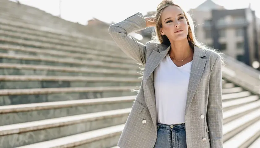 Chiara Ferragni w szarej marynarce w kratę i jeansach, z ręką we włosach, na tle schodów.
