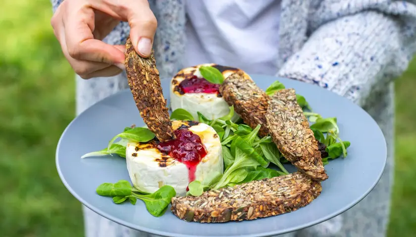 Ręka zanurza chrupiący sucharek w grillowanym serze z konfiturą. Czy takie sucharki są kaloryczne? Zależy od składu, ale te pełne ziaren wyglądają zdrowo!
