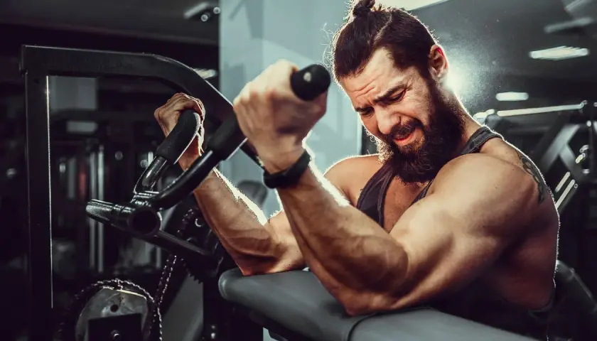 Mężczyzna z brodą i kitką ćwiczy biceps na maszynie. Widać jego napięte mięśnie i determinację. To idealne miejsce, by budować **mutant mass sklad**.