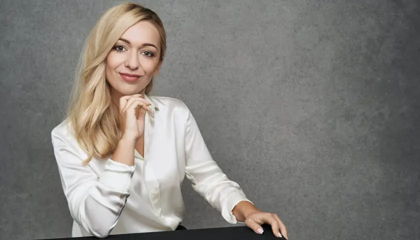 Blondynka w białej satynowej koszuli podczas sesji modowej, z dłonią pod brodą, uśmiecha się do obiektywu.