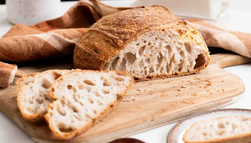 Un delicioso pan de masa madre, con su corteza dorada y miga aireada, reposa sobre una tabla de madera junto a unas rebanadas.