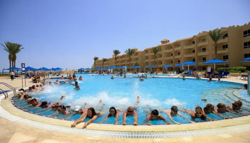 Grupa ludzi pływa w basenie hotelu, tworząc falę. W tle budynek hotelu i palmy. Wiele pozytywnych amwaj blue beach resort & spa opinie potwierdza ten widok.