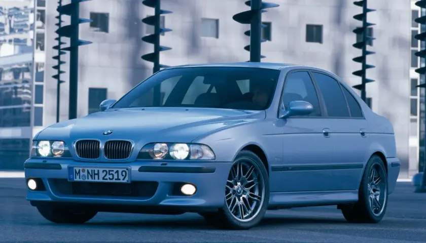 Błękitne e39 bmw z felgami M, zaparkowane przed nowoczesnym budynkiem.