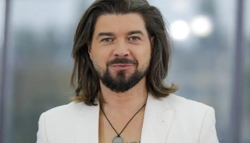 Tomasz Szczepanik w białej marynarce, z długimi włosami i brodą, patrzy w obiektyw.