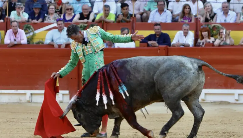 Un torero alicantino, con traje de luces verde, se enfrenta a un toro en la arena. El público observa desde las gradas.