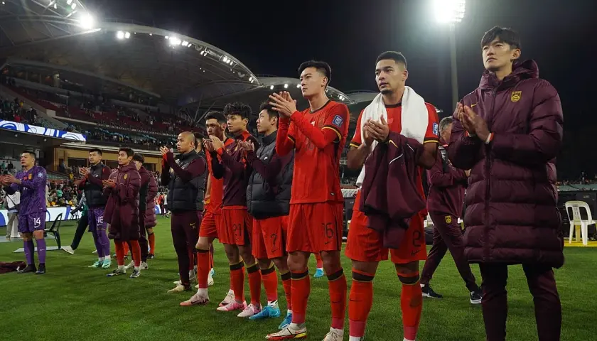 Piłkarze w czerwonych strojach, być może reprezentacja Chin, klaszczą na boisku. W tle widać trybuny i światła stadionu.