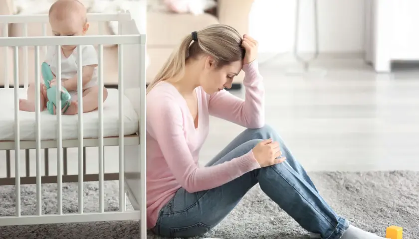 Zmęczona mama siedzi na podłodze obok łóżeczka, czując baby blues. Dziecko bawi się zabawką.