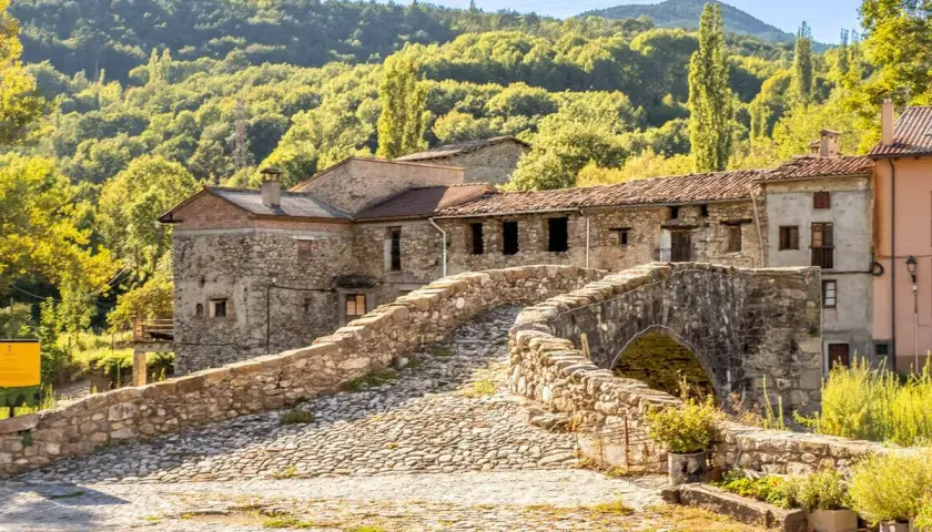 Puente de piedra y casas rústicas en el Pirineo de Lleida, rodeado de bosques verdes bajo un cielo azul.