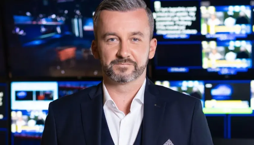 Krzysztof Skórzyński, elegancki mężczyzna w garniturze, stoi na tle ekranów. Ile ma lat? Tego nie wiemy, ale wygląda na doświadczonego dziennikarza.