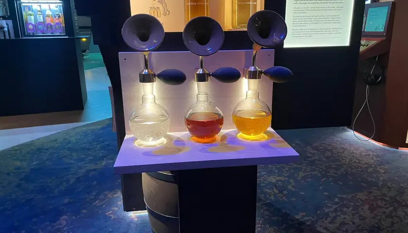 Tre flaskor med olika färger av sprit står upplysta på en hylla. En utställning på Vin & Sprit Museum.