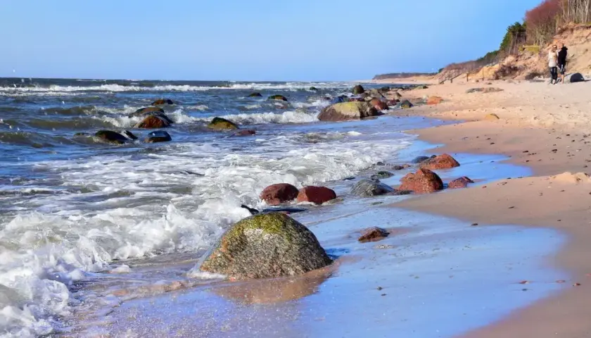 Kamienista plaża w Dębinie, z falami rozbijającymi się o brzeg i dwiema osobami spacerującymi po piasku. Dębina atrakcje przyrodnicze zachwycają.