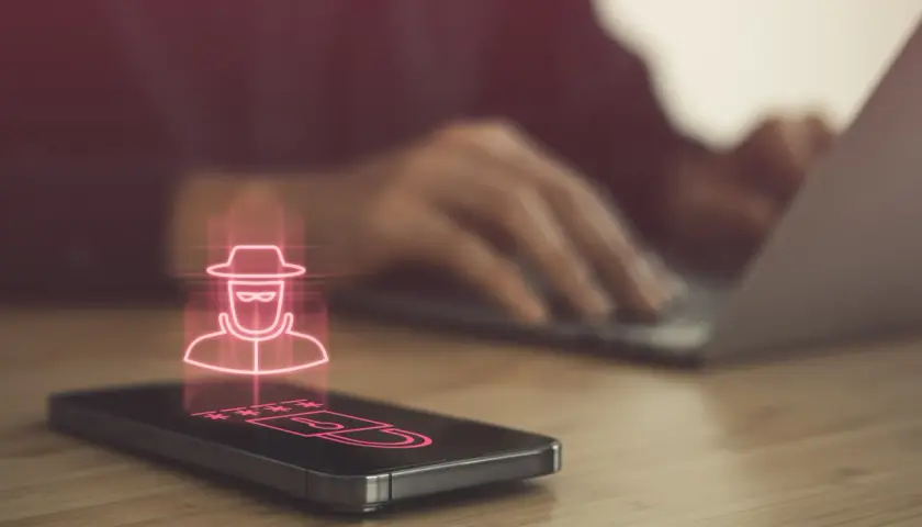 Haker w kapeluszu i gwiezdki symbolizujące hasło na ekranie telefonu, sugerujące podsłuch w telefonie. W tle osoba pracuje na laptopie.