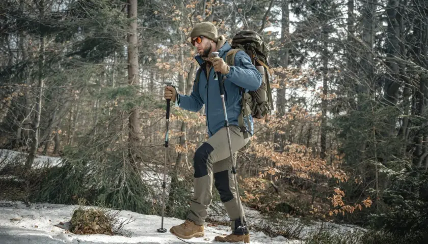 Mężczyzna w zimowej kurtce i czapce idzie przez las, wspierając się na kijach trekkingowych. W tle drzewa i śnieg.