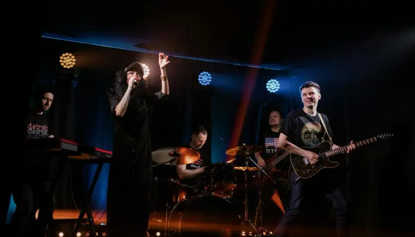Energiczny zz trio rockowe na scenie. Wokalistka w czarnej sukience, gitarzysta, basista, perkusista i klawiszowiec tworzą dynamiczny zespół.