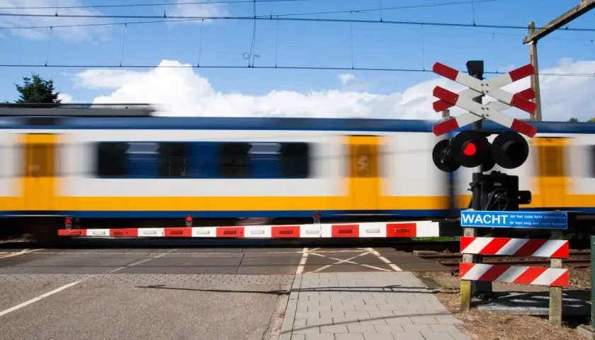 Pędzący pociąg mija przejazd kolejowy z czerwonym światłem i znakiem "Wacht". Widoczny jest charakterystyczny krzyż Andrzeja.