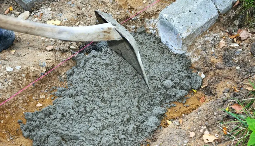 En hacka sprider jordfuktig betong i en grop. Betongblock ligger bredvid.