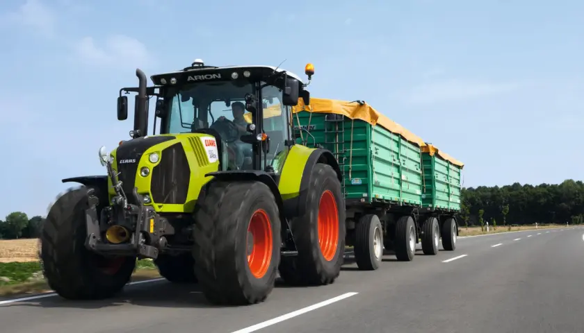 Ein Claas Arion Traktor mit Anhängern fährt auf der Straße. Der Fahrer hat die Fahrerlaubnis L und transportiert landwirtschaftliche Güter.