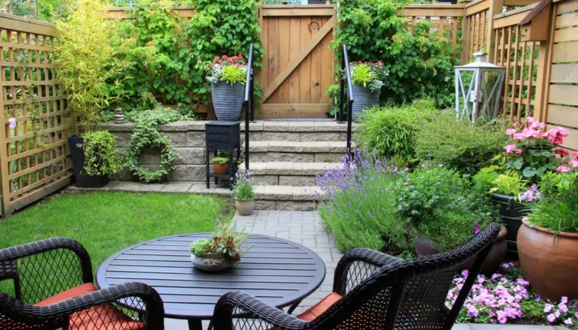 Ein kleiner Garten mit Sitzgruppe, Stufen und Holztor. Üppige Pflanzen und Blumen schaffen eine grüne Oase.