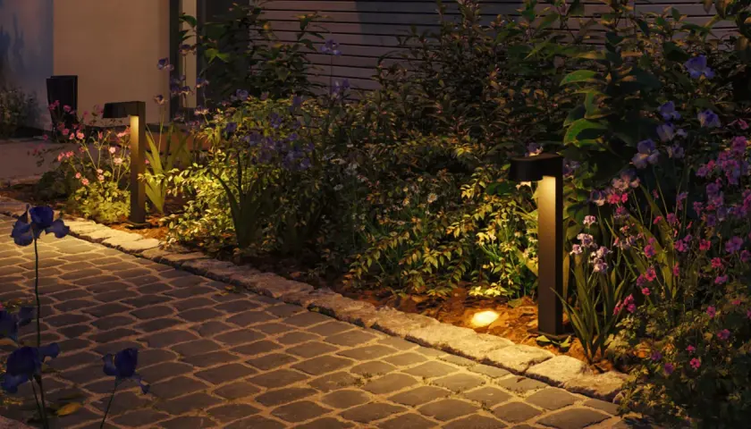 Beleuchteter Gartenweg mit Blumen und Büschen. Das warme Licht der Gartenlampen zieht kleine Insekten an, die im Schein tanzen.