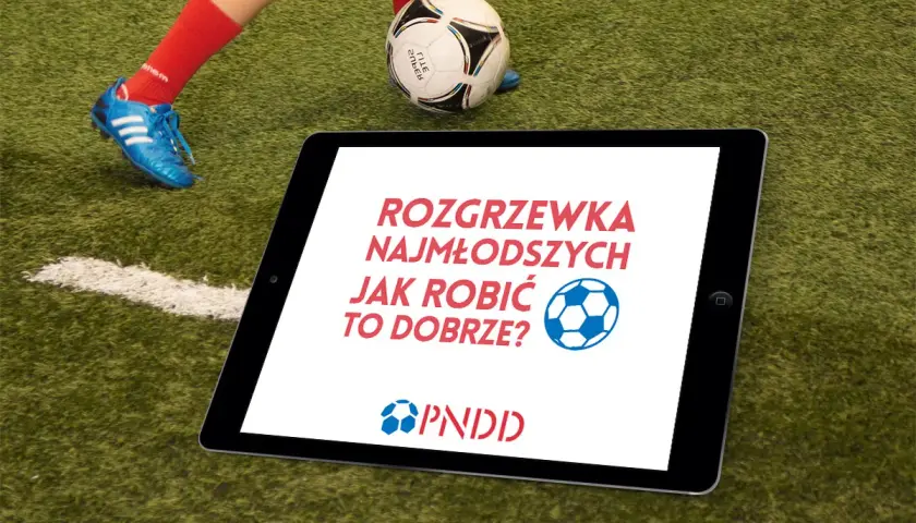 Tablet na boisku z napisem "Rozgrzewka najmłodszych jak robić to dobrze?". Obok piłka i noga dziecka w czerwonej skarpecie.
