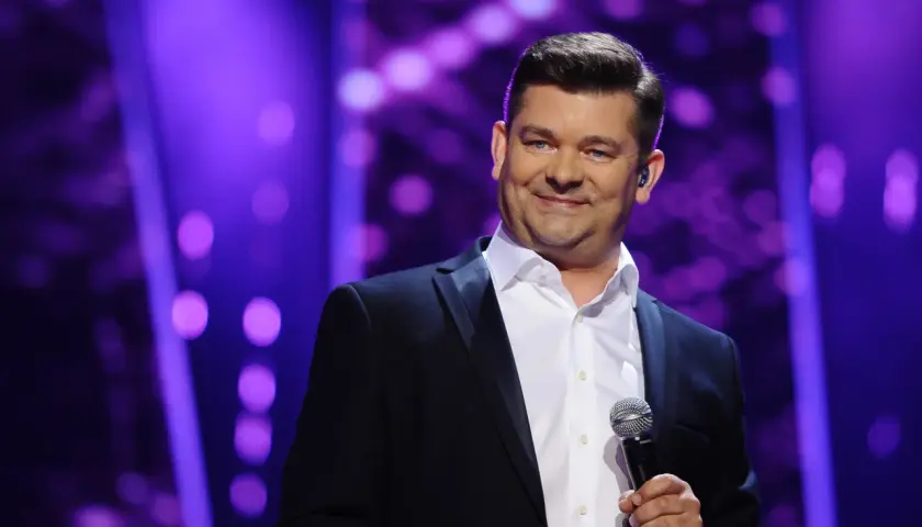 Zenek Martyniuk w czarnym garniturze i białej koszuli trzyma mikrofon na tle fioletowych świateł.