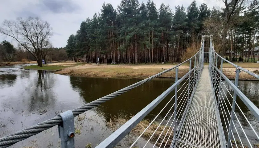 Metalowy most wiszący w Polsce nad rzeką, otoczony lasem sosnowym i piaszczystym brzegiem.